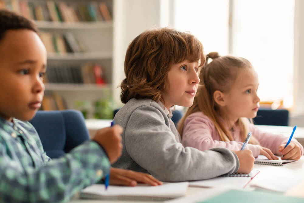 Elementary classroom of diverse children listening attentively to their teacher, writing in exercise notebooks